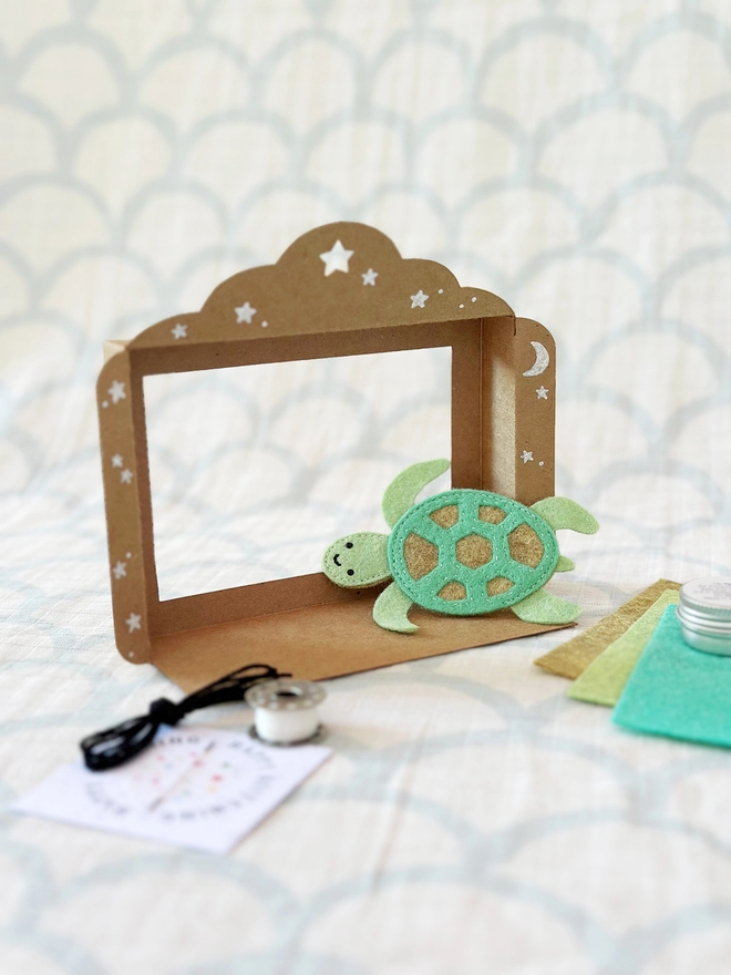 A handmade mini cardboard puppet theatre stands on a fabric surface, with a sea turtle finger puppet beside it.