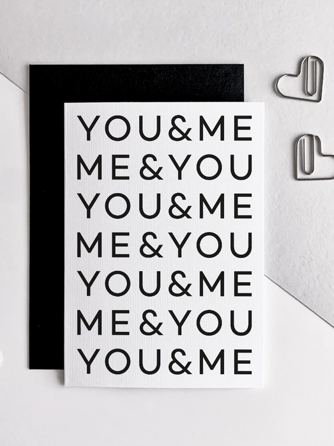 You & Me Anniversary Card overlapping a black envelope next to heart shaped paperclips.