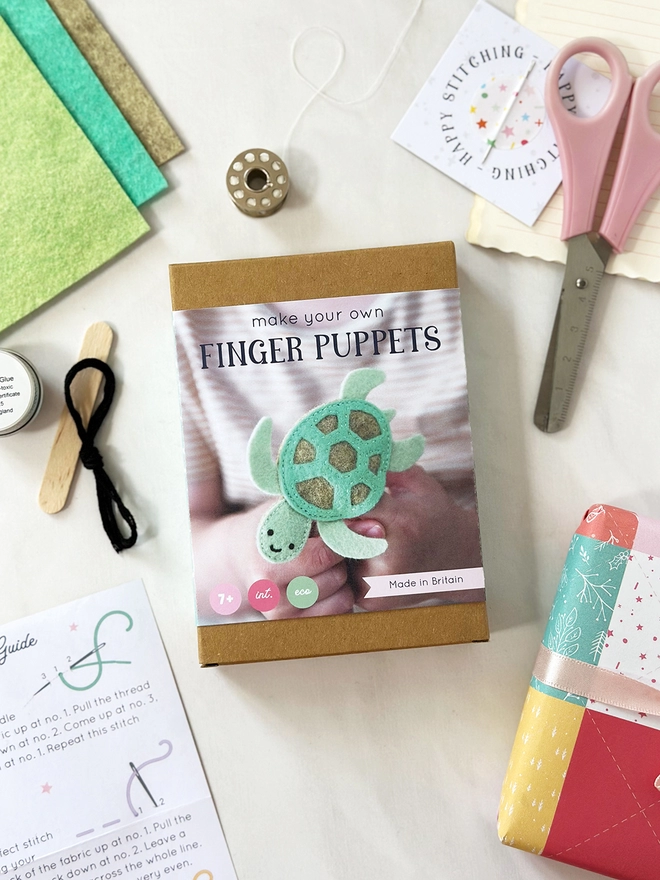 A small craft kit box to make a felt sea turtle finger puppet, lays on a white desk. Various craft kit contents lay around the box.