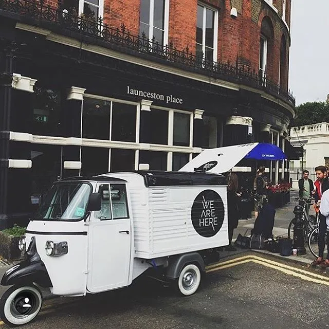 A black and white mobile coffee tuk tuk with We Are Here on the side in a big black circle, the boot is open at the rear to make coffees for a film crew outside of a pub in London
