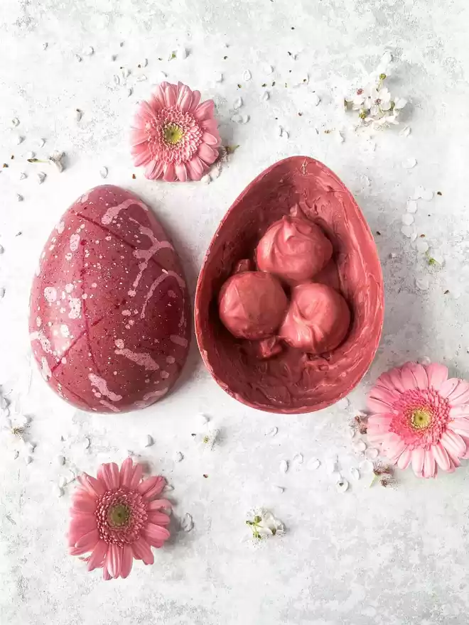 A pink easter egg sitting on top of a white background with a half open egg showing chocolate covered macarons embedded inside