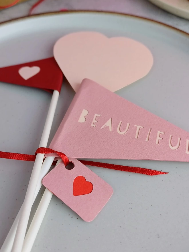 A closeup of 3 decorations laid on a plate tied together with a tiny pink gift label. The decorations include a cherry red paper flag with pale pink heart, a pale pink heart decoration and a blush pink flag with the word beautiful in pale pink.