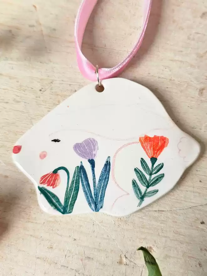 flat ceramic ornament shaped like a bunny with flowers hand painted on it, ears eyes and a nose. hanging with pink velvet ribbon