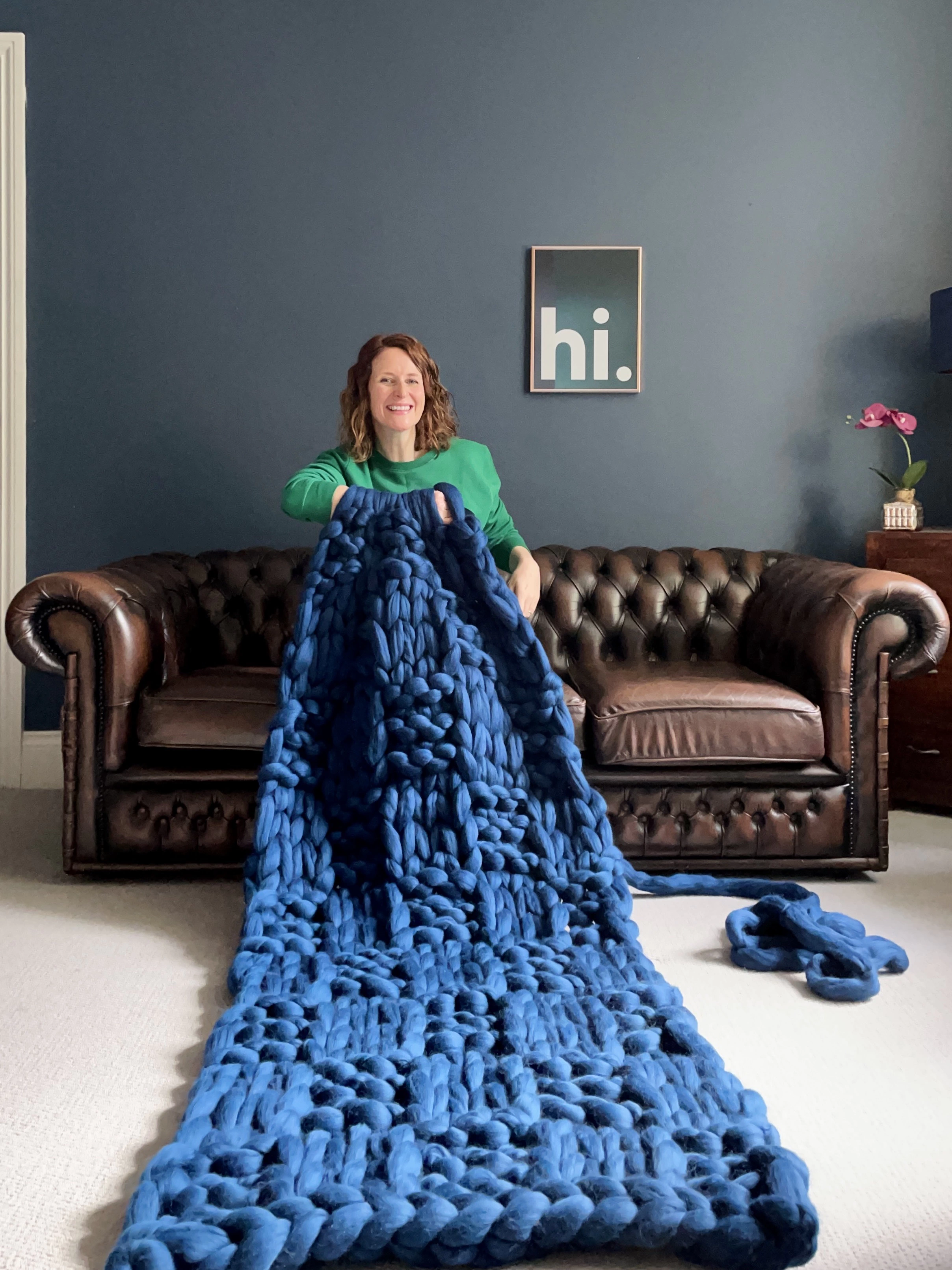 Mizz, a white woman wearing a bright green jumper, sits on a brown leather sofa in front of a blue wall with a framed print that says "hi". She is arm knitting a huge rich ocean blue blanket, and the bumps of the knit and purl stitches reach out towards you. 