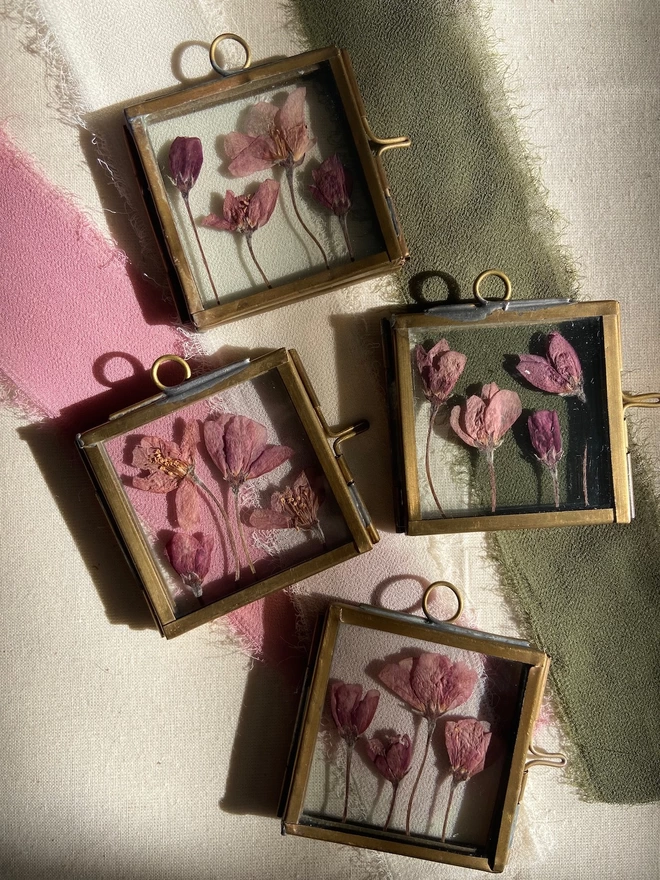 Four square brass frames with small pink pressed flowers laid over chiffon ribbon