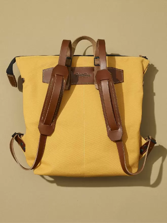 Back view of yellow zip top backpack with brown leather trim on taupe background.