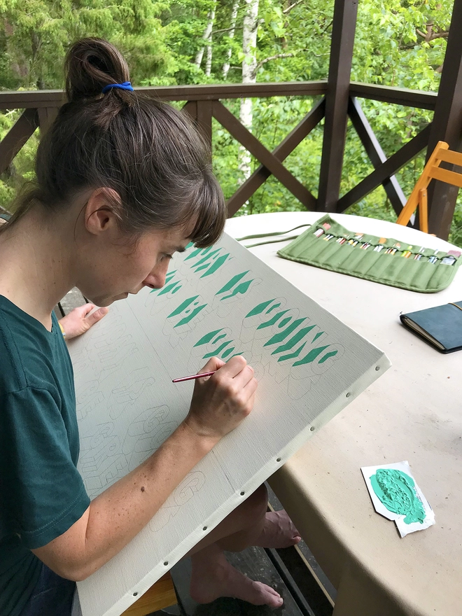 Artist Survival Techniques painting green onto a canvas that’s leaning on a table with lush green trees in the background. 