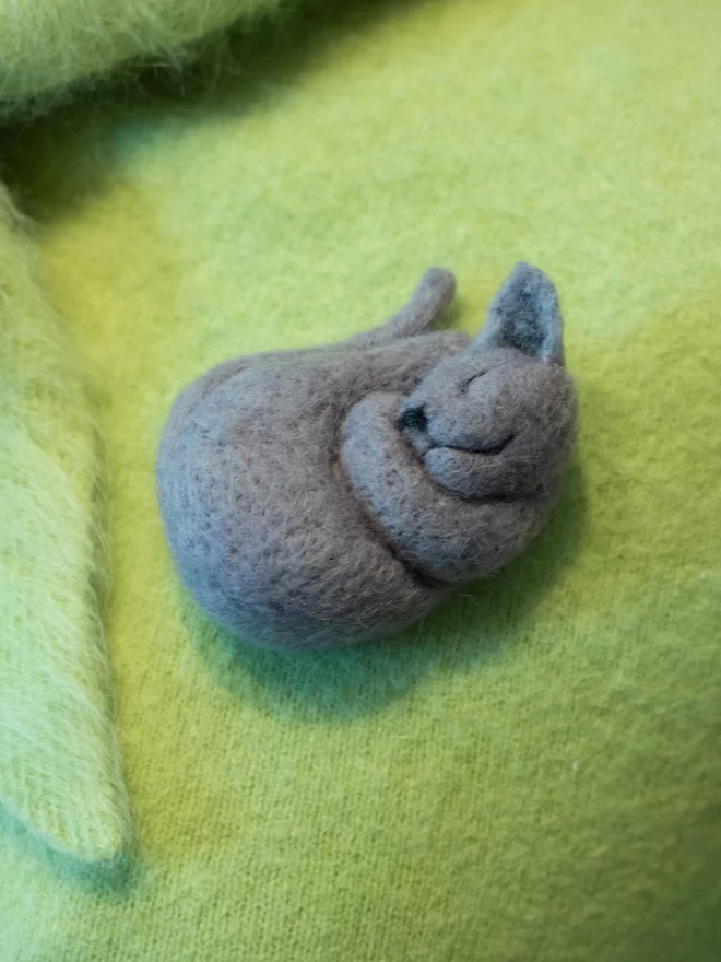 A needle felted grey sleeping cat brooch on a green fluffy jumper 