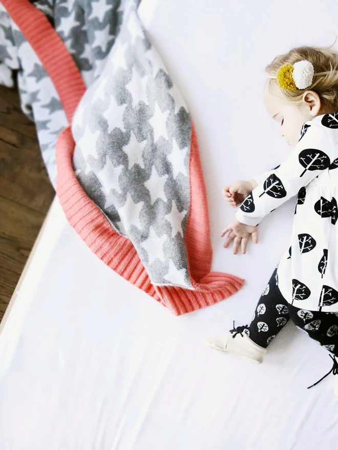 A toddler lies asleep on a bed. A grey and white junior star blanket with coral pink trim is draped half on and half off the bed.