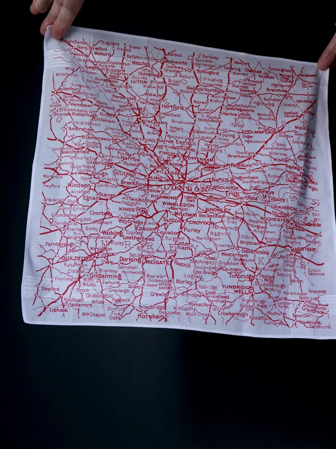 A Mr.PS Not Far From London map hankie printed in red held aloft by two hands in front of a dark wall 