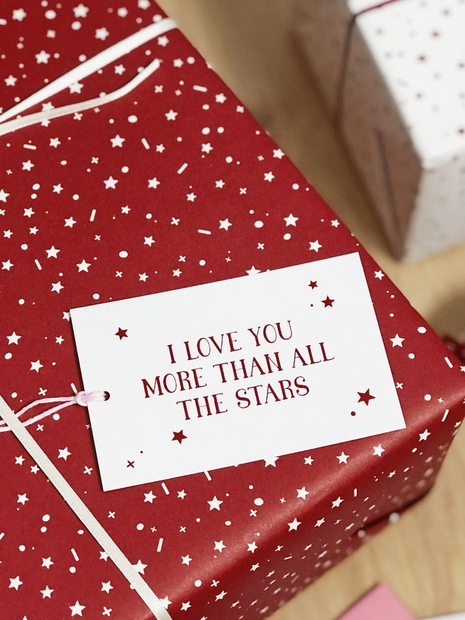 A gift wrapped in a red star design wrapping paper and a tag that reads "I love you more than all the stars" is on a wooden surface.