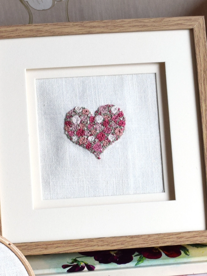 An embroidered Pink Roses Heart, of woven wheel roses in 5 shades of pink with French Knot green grass.  Displayed in an oak frame.