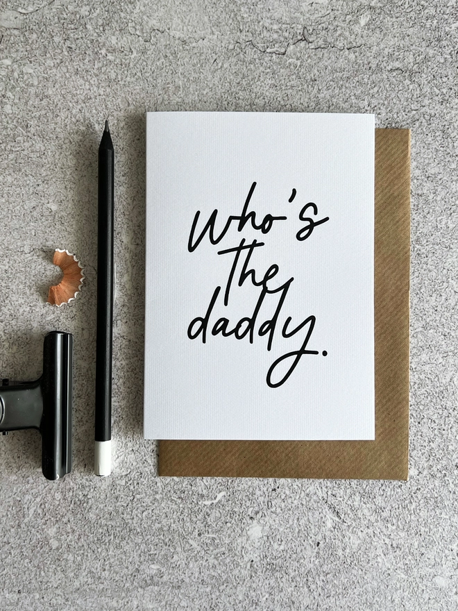 White card that reads "Who's The Daddy" in black cursive, laid on a brown envelope, next to black items of stationary.