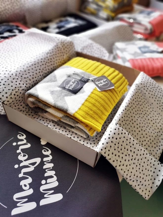 A neatly folded chevron blanket is shown packed inside a box and wrapped with tissue paper. The box is sitting on a table surrounded by other gift wrapped products. Beneath the box is a grey Marjorie Minnie branded gift bag.
