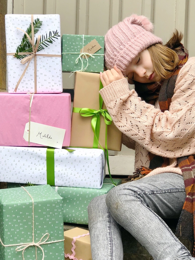 A stack of gifts wrapped in green and white paper with a tiny tree design are piled beside a sleeping girl.