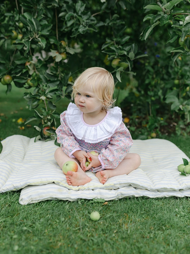 A little girl in a floral smocked romper with a white frilled collar sits on a mattress under a tree