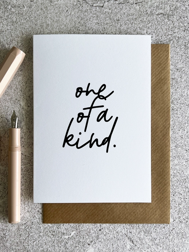 One Of A Kind Thank You Card that reads "one of a kind" in cursive, laid on a brown envelope next to a pale pink pen.
