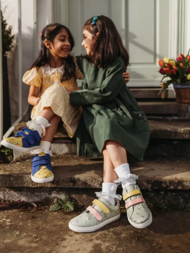 Two girls sitting on a step laughing wearing pip and henry double Velcro strap trainers