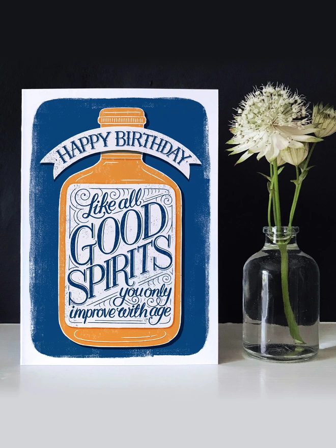 birthday card with hand lettered label saying like all good spirits you only improve with age on a dark background with white flower