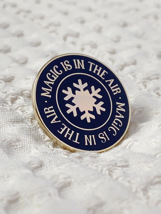 A navy blue and gold enamel pin badge with a gold star in the centre and the words "Magic is in the air" around the outside is on white fabric.