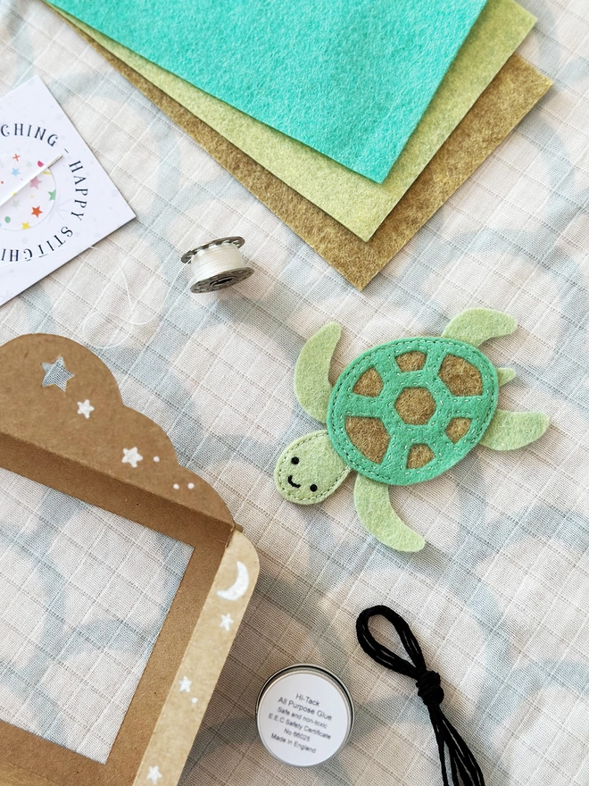 A handmade felt sea turtle finger puppet is surrounded by the craft kit contents used to make it.