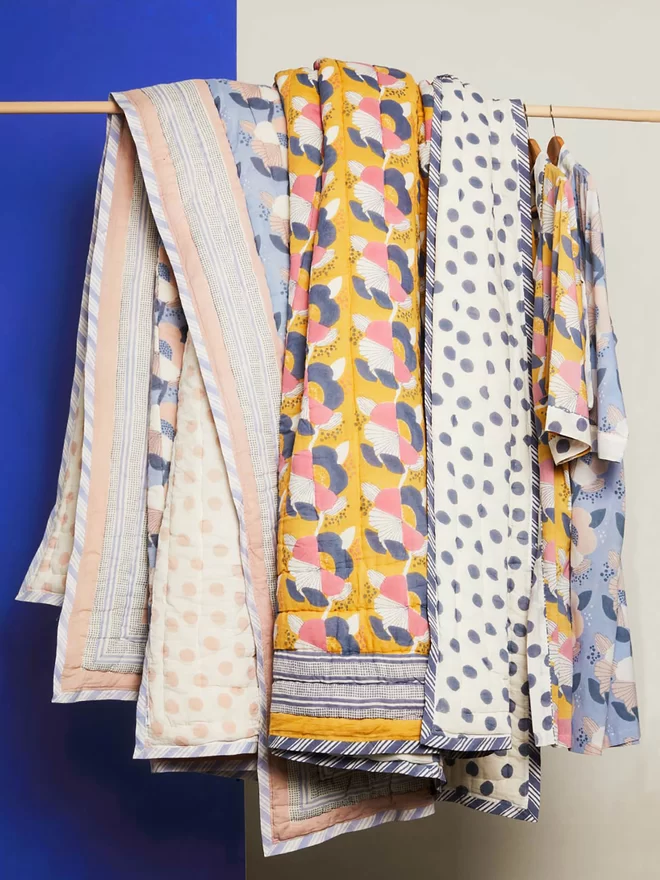 Collection of block printed quilts in blue and yellow tones hanging over wooden pole in front of a bright blue background