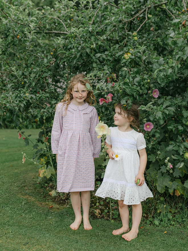 Two girls stand under a tree. One is wearing a pink long-sleeved dress with a collar. The other is in a white dress.