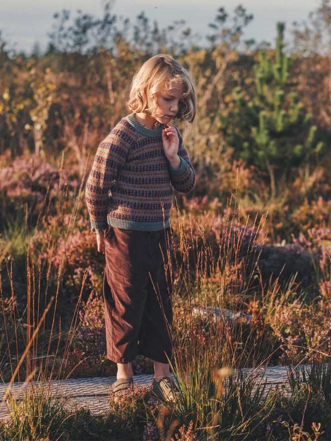 A boy stands in the middle of a bog strewn in heather wearing a moss green fairisle jumper and brown trousers