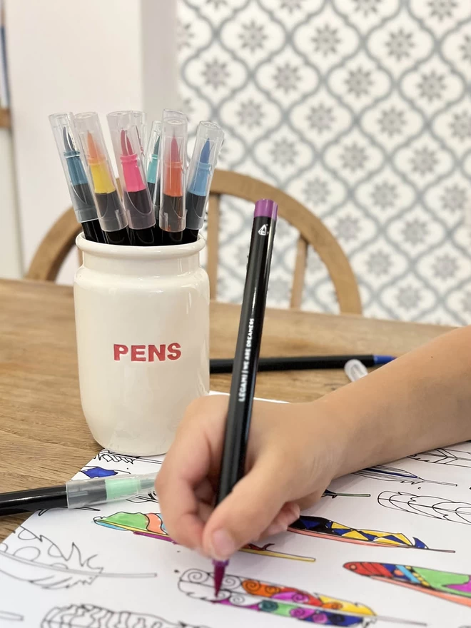 A child is colouring a picture, next to a handmade pens pot full of brightly coloured felt tips.