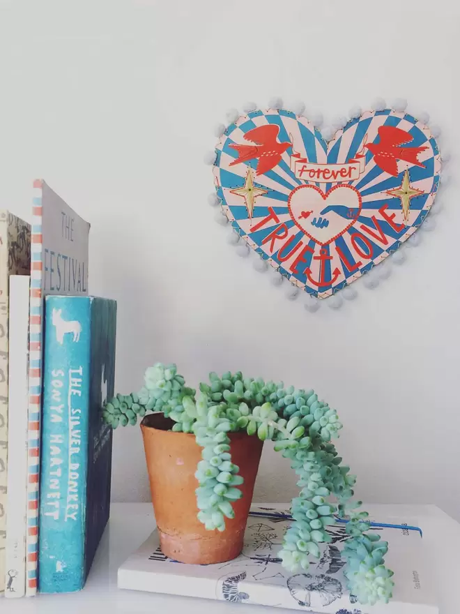 The colourful wooden love heart is show hung on the wall near some books and a plant. 