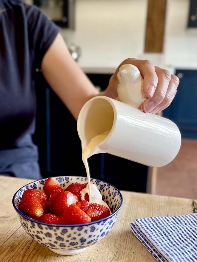 A handmade ceramic bird jug is being used to pour cream over a bowl full of strawberries. 