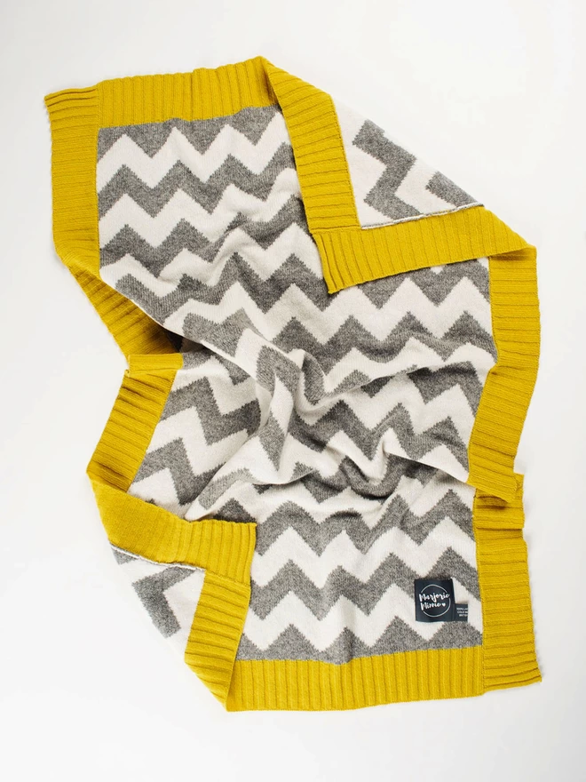 A flatlay of a knitted baby blanket with grey and white chevron pattern and mustard yellow ribbed trim. The corners are slightly ruffled and turned back to reveal the reverse colourway.