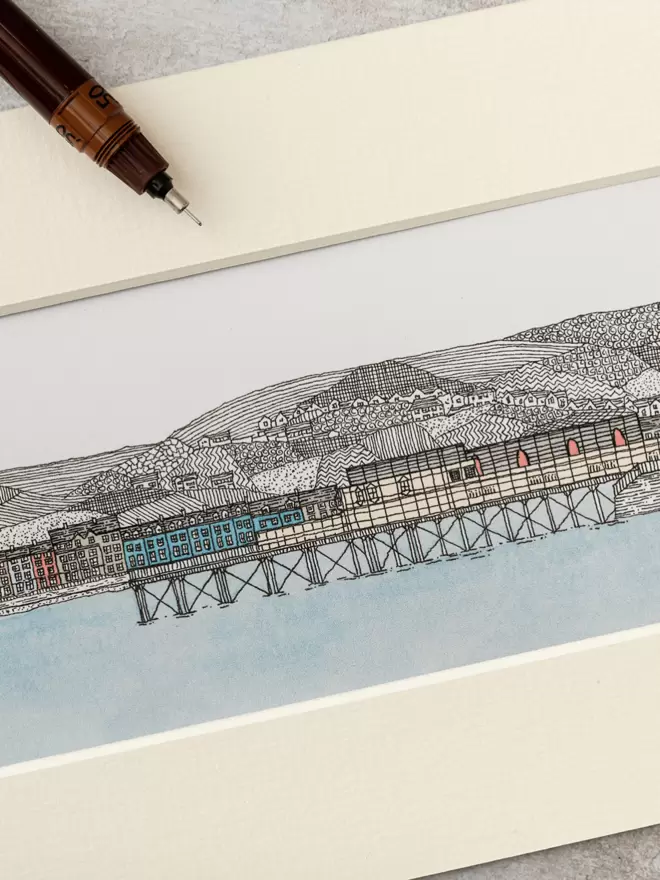 Print of detailed pen and watercolour drawing of Aberystwyth pier and seafront, with mountain backdrop, in a soft white mount