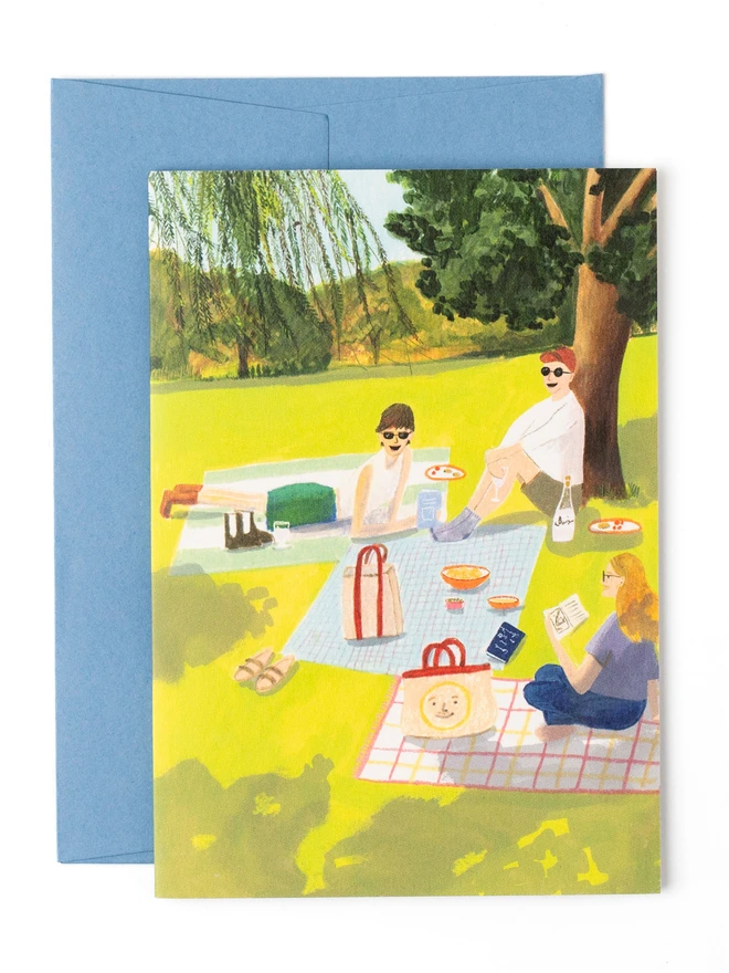A greeting card featuring a summery illustration of three friends having a picnic in the dappled light under a tree. They are drinking wine and eating olives whilst chatting