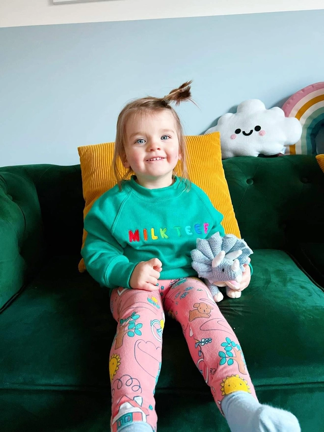 A young child sitting on a green sofa wearing a rainbow embroidery 'Milk Teef' slogan sweatshirt.
