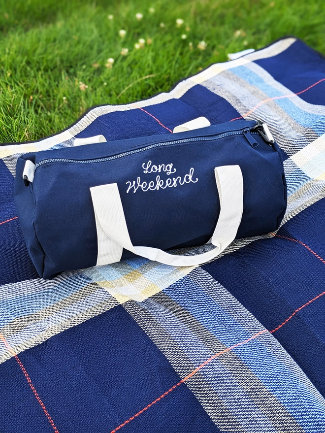 Navy Blue Duffel Bag with embroidered 'Long Weekend' sitting on a blue checkered picnic blanket in a field on a summers day