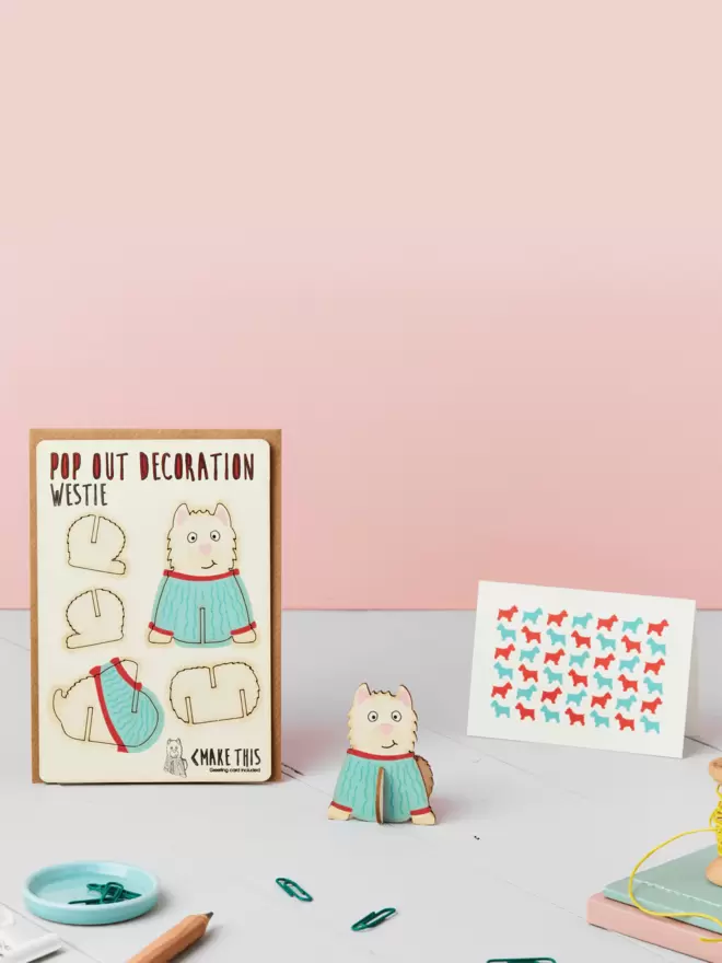 3D westie dog decoration and westie pattern greeting card and brown kraft envelope on top of a grey desk in front of a light pink coloured background