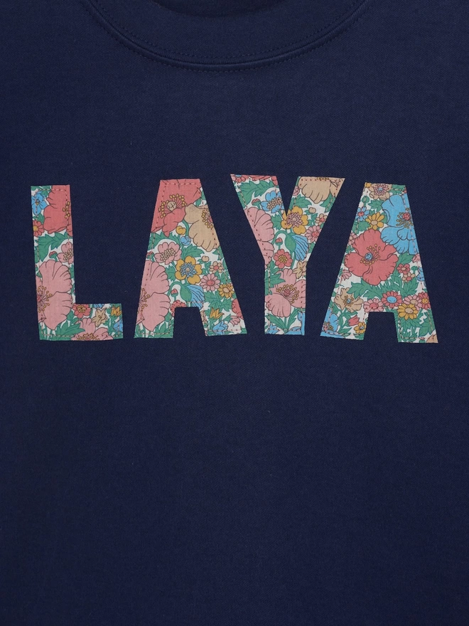  a navy t-shirt with the name Laya appliquéd on the fron Floral Liberty Print t , close up on the stitching 