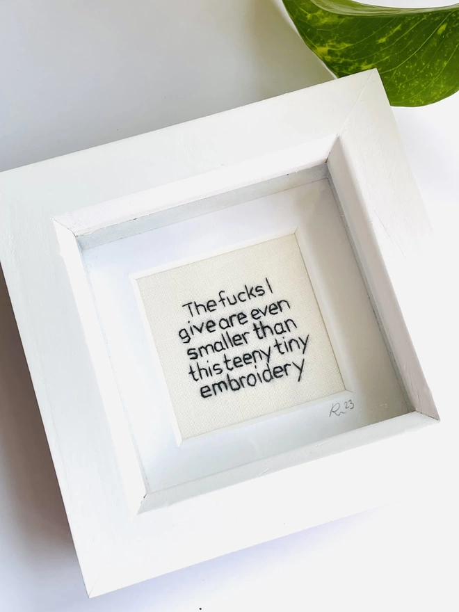 A miniature white box frame with hand embroidered black text that reads The fucks I give are even smaller than this teeny tiny embroidery