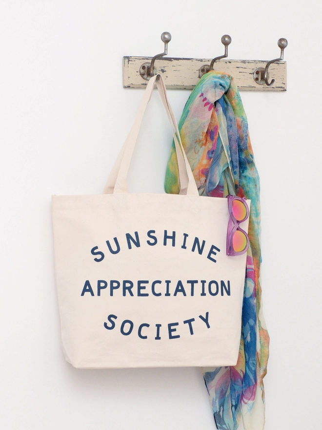 a large natural canvas tote bag bearing the words sunshine appreciation society hanging on a hook