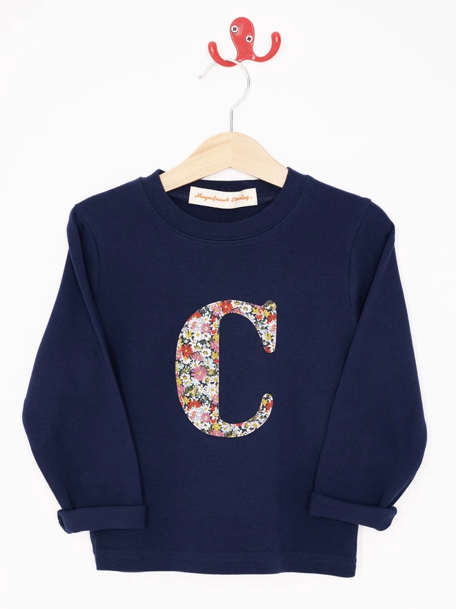 A navy cotton long sleeve t-shirt appliquéd with an initial in a floral Liberty print, hanging on a hanger 