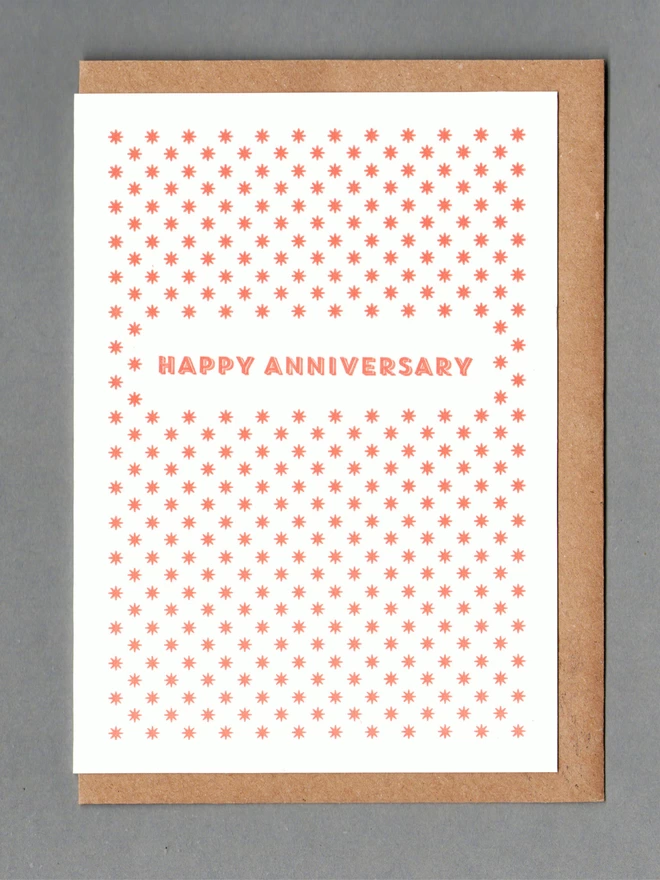 White card with orange stars and orange text reading 'HAPPY ANNIVERSARY' with a kraft envelope behind it