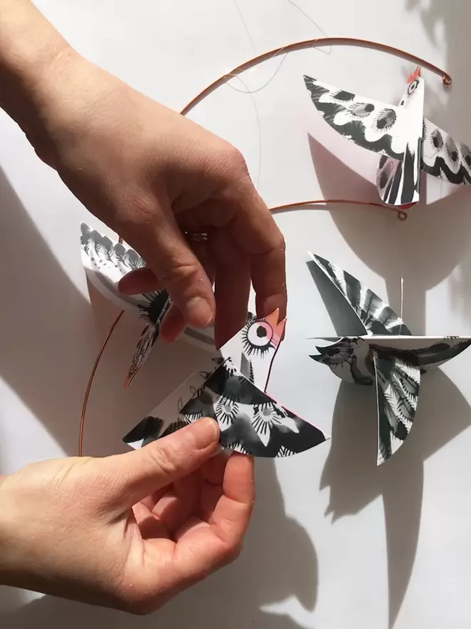 Artists' hands hold a folded black and white paper bird, with 3 other birds in background