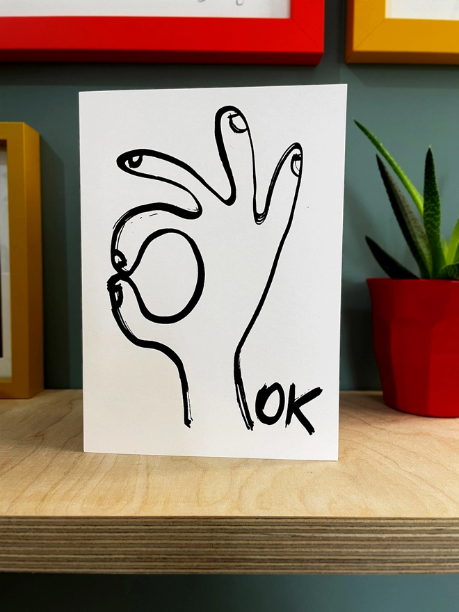 A white Ok Greetings Card with a black ink drawn hand and the word ok, sits on a plywood shelf with glimpse of picture frames around and a plant to one side.