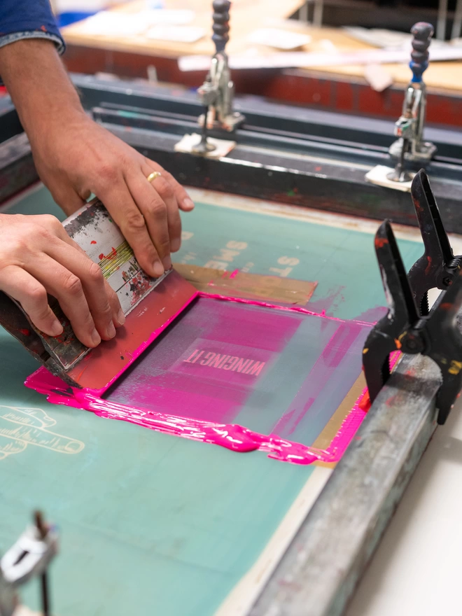 Hands holding a squeegee, pulling flouro ink across a screen printing screen 