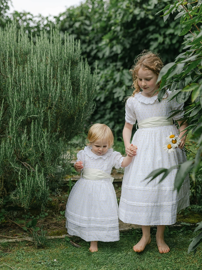 A big and little girl in matching white dresses with yellow sashes are holding hands in a garden