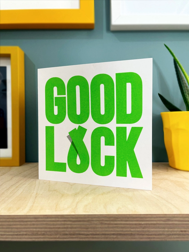  BOLD green lettering spells out good luck on this square card, with the U of luck as a crossed finger. Simple, timeless and in lucky green, sits on a plywood shelf with a few picture frames around the edges and a pot plant in a yellow cup.