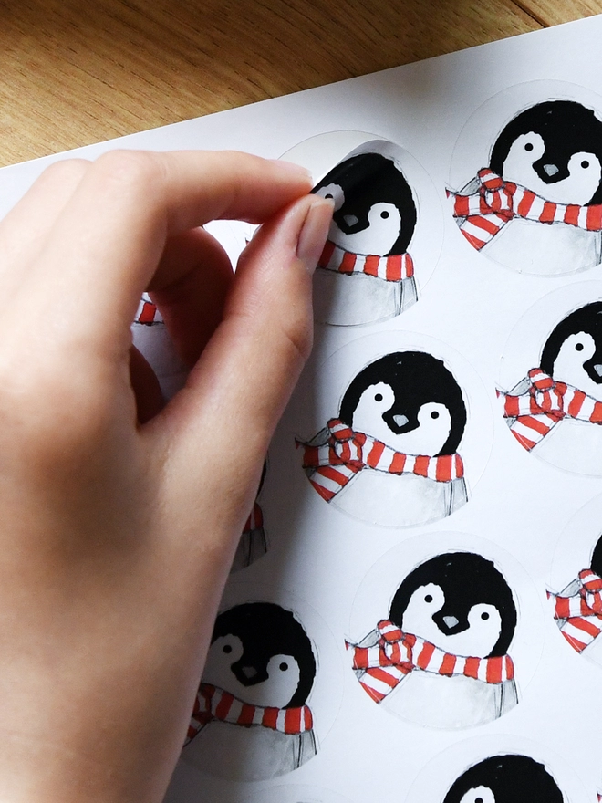 A hand is peeling a round sticker with a baby penguin design off a sheet of 35 stickers.