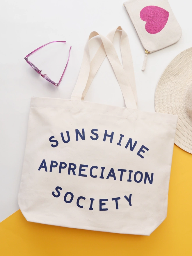 a large natural canvas tote bag bearing the words sunshine appreciation society laying on a white and yellow background next to a sunhat, sunglasses and purse