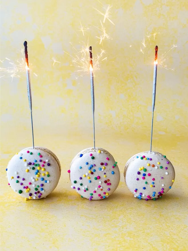 three macarons adorned with colourful sprinklers. Each macaron has a sparkler on top of them. All against a yellow background
